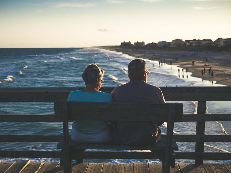 Older woman and older man sitting on a bench on a boardwalk overlooking the ocean.
