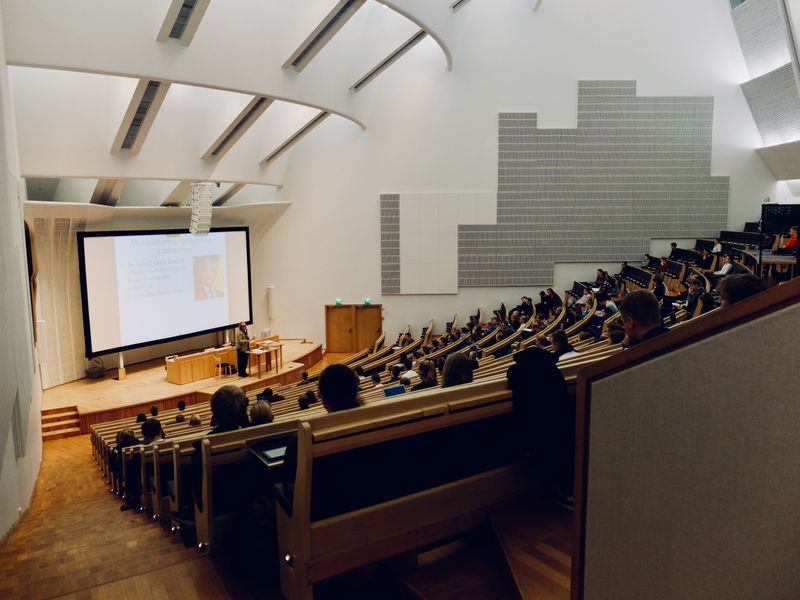 A teacher is presenting in front of a large screen in a lecture hall that is half full of students.  