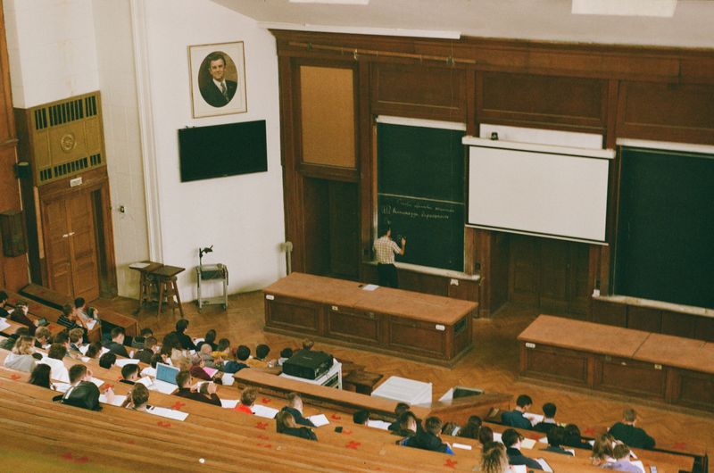 A university classroom with many students listening to a teacher as he lectures and writes on a chalkboard.