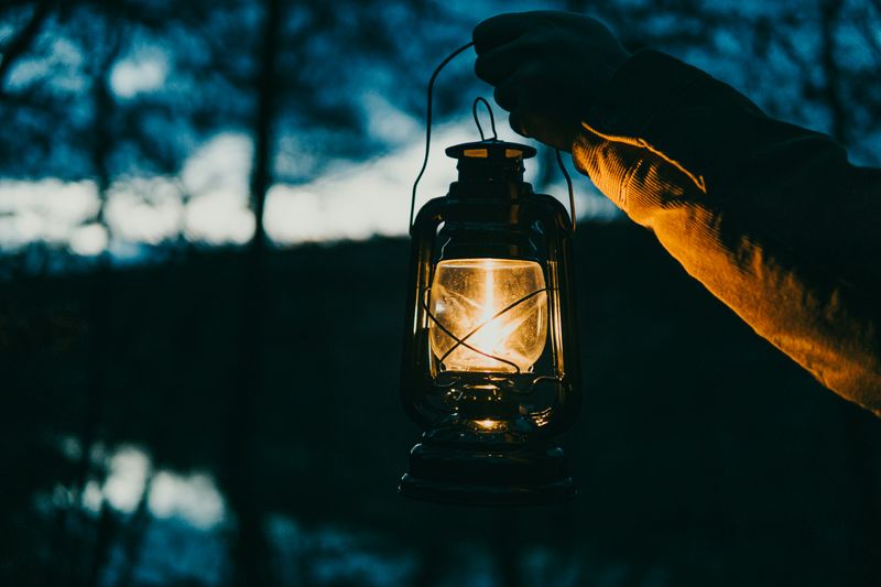 Someone wearing a sweatshirt holding a lit, black lantern outside while it is getting dark.