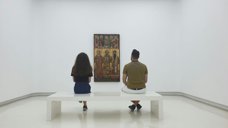 Two people sitting on a bench in front of a large painting in a gallery.