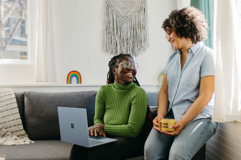 Image: A couple sitting together making eye contact and smiling, one with a laptop, while the other holds a mug.