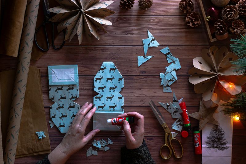 A person using a glue stick on a handmade paper ornament with scissors, glue sticks, pine cones, and paper in the background.