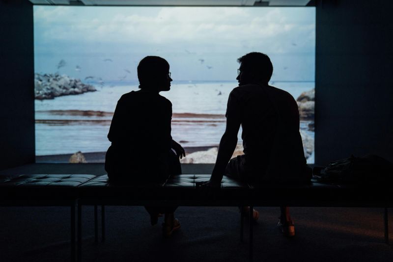 Two people on a bench, silhouetted against a large screen or window that displays a seascape with gentle waves.