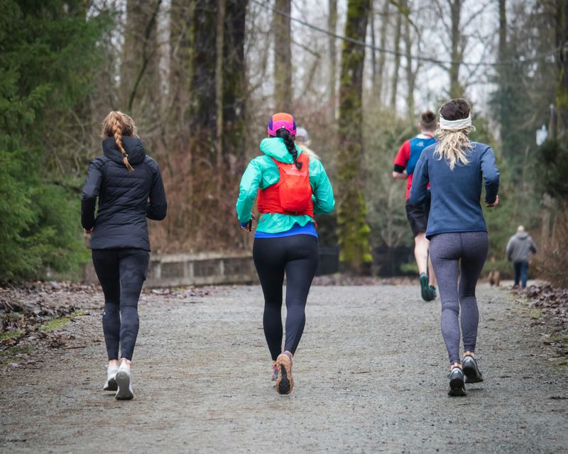 Three women run side by side on a tree‑lined gravel path, with more runners visible ahead of them.