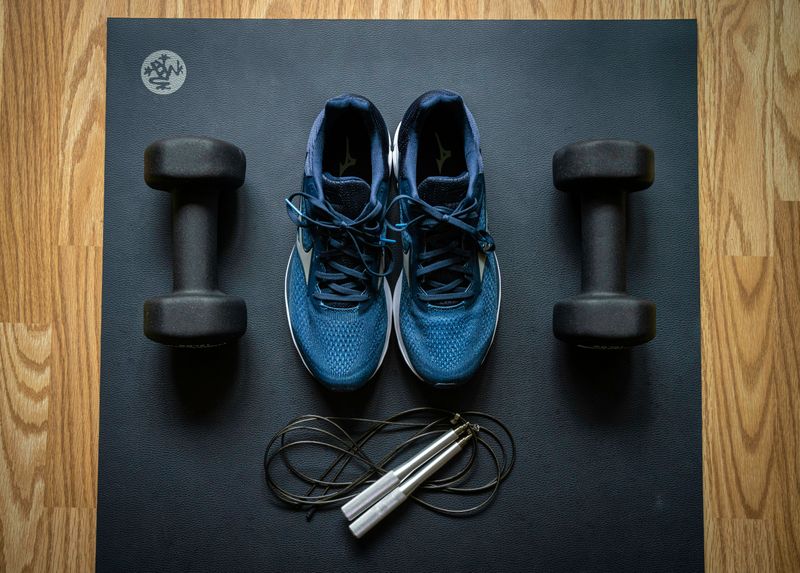 A yoga mat with weights, running shoes, and a skipping rope laid out on the floor.