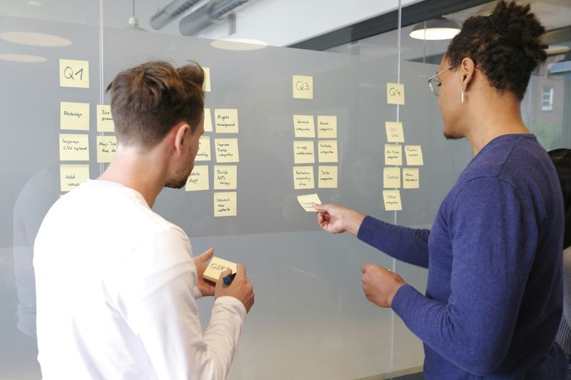 Two people facing a glass wall with sticky notes, divided into "Q1," "Q2", "Q3", and "Q4".