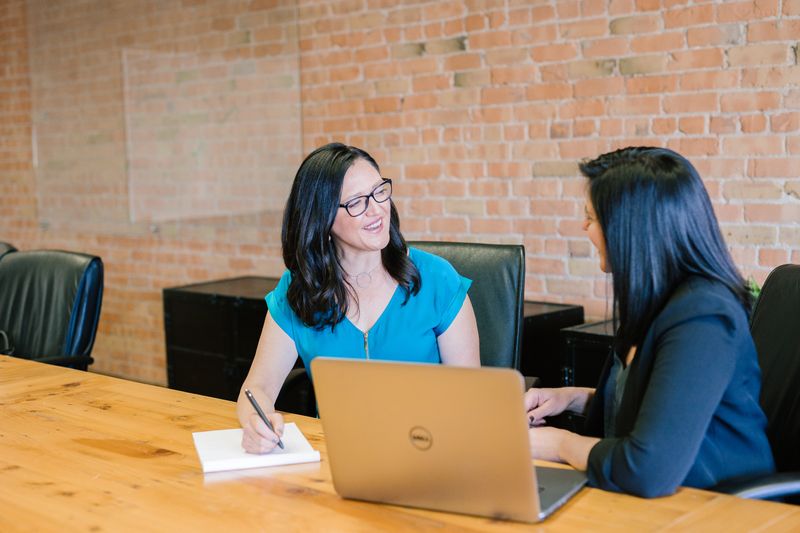 Two dark haired women seated beside one another. One is holding a pen and writing on a white notepad; the other has a laptop.