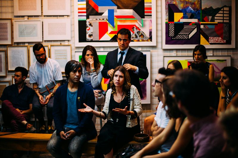 A room full of people. One woman in the center of the room is talking and the others are listening to her. 