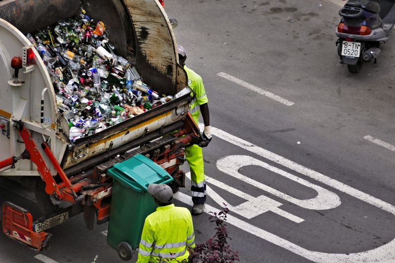 A city street with a garbage truck filled with trash and two sanitation workers.