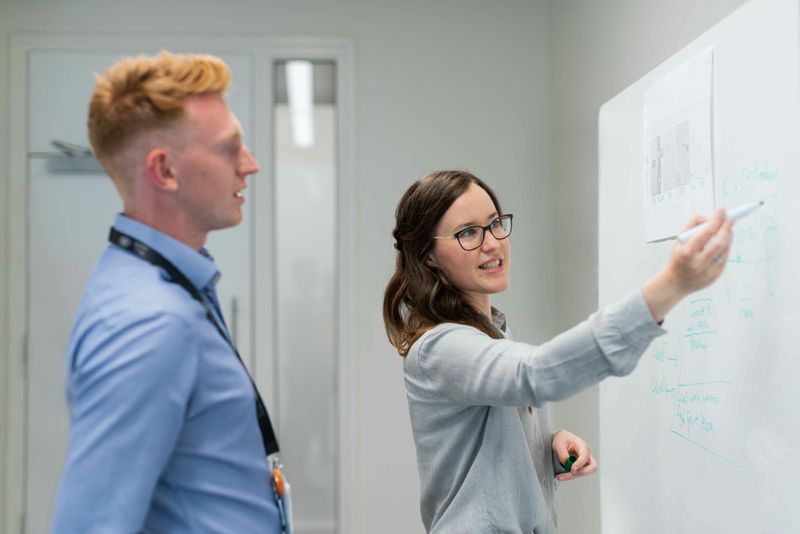 A woman writes something on the whiteboard while a man looks at the board. 