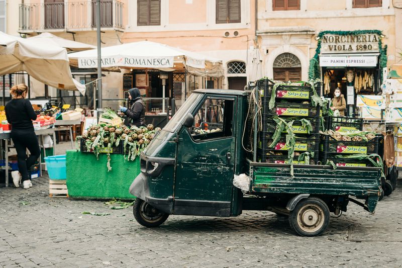 A truck with crates of fresh food in the back driving through an Italian city.