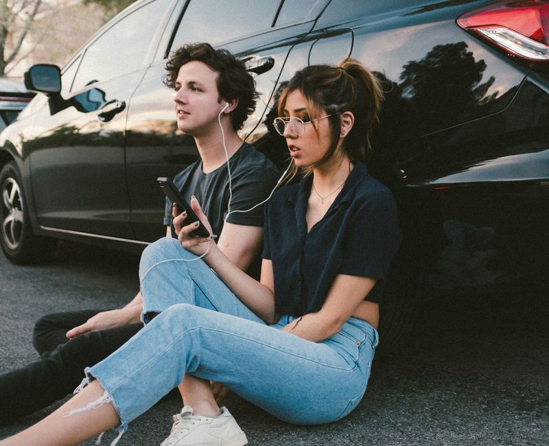 A man and a women, sitting against a car, sharing earphones, listening to something on a phone.