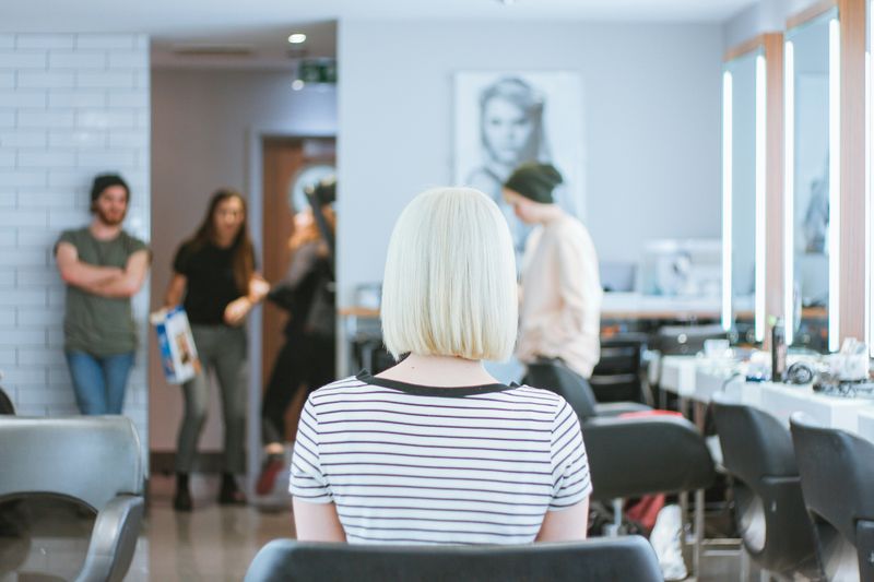 People in a barbershop. A blonde woman sits on a barber's chair.