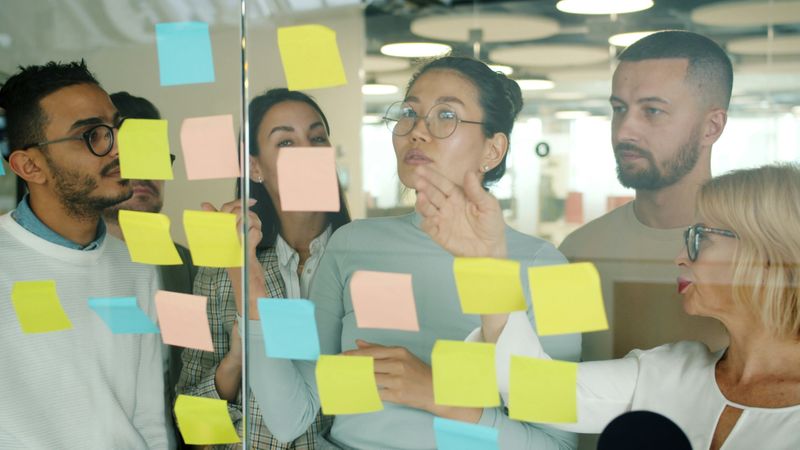 A group of coworkers look at different colored sticky notes on a glass wall.