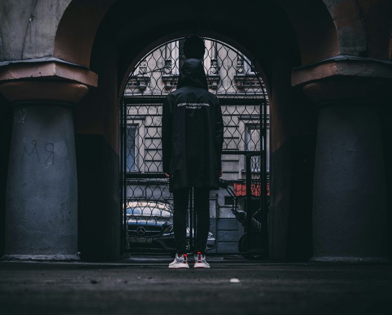 A person in all black stands in front of a closed iron gate outside of a university.