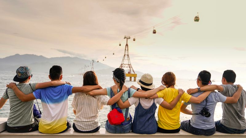 Eight friends sitting on the edge of pier holding shoulders and looking at gondolas crossing a river gorge.