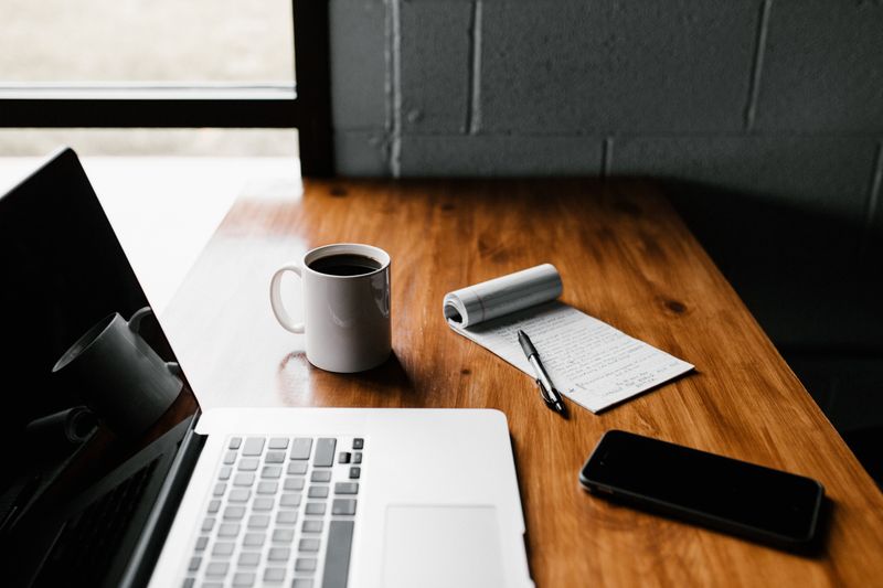 A laptop on a desk beside a coffee cup, a phone, and a notepad