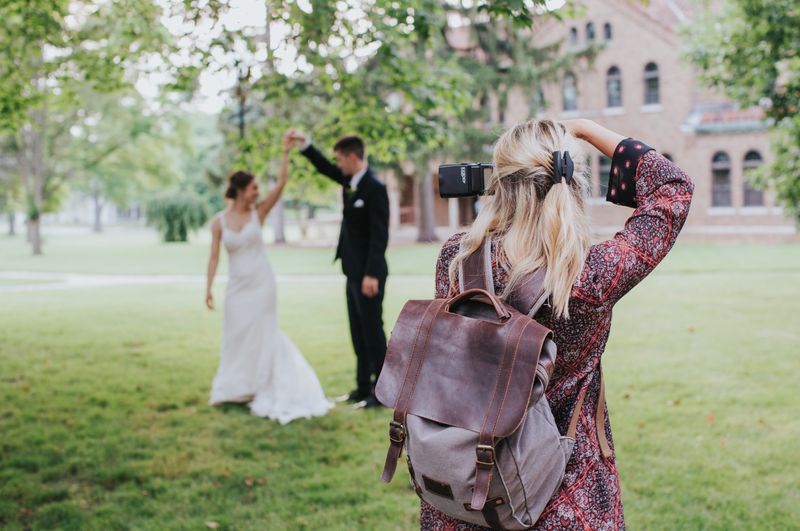 A wedding photographer taking picture of the bride and groom in a field under a tree.
