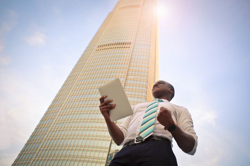 Young man with white button up shirt, black pants, and striped teal tie standing in front of a tall office building