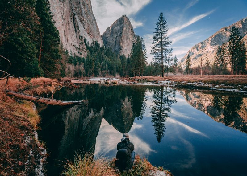 Person with a backpack in the foreground is gazing on a beautiful nature scene of mountains & trees reflecting in the water. 