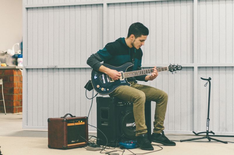 A man playing the electric guitar in a rehearsal space while sitting on an amplifier.