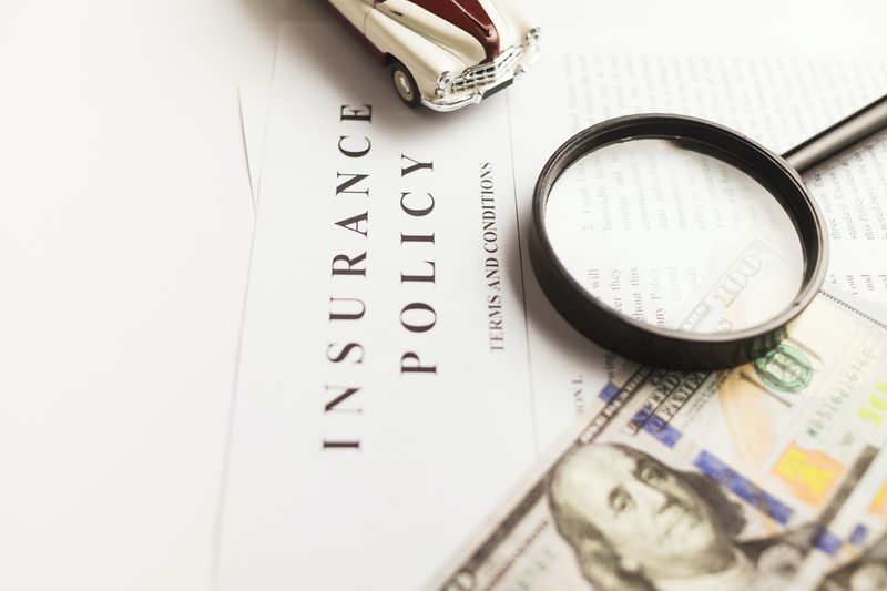 An image of an insurance policy. On top of the policy is a toy car, magnifying class, and a one hundred U.S. dollar bill.