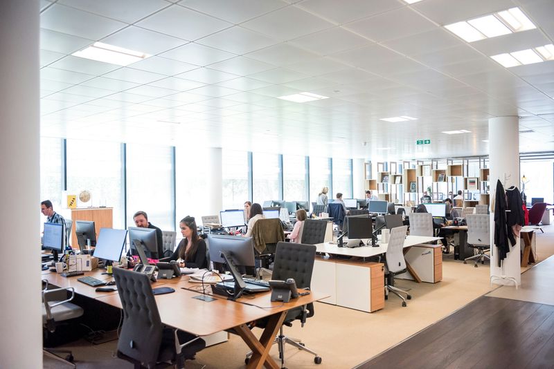 Open office layout with people working on computers at large shared desks in a bright room