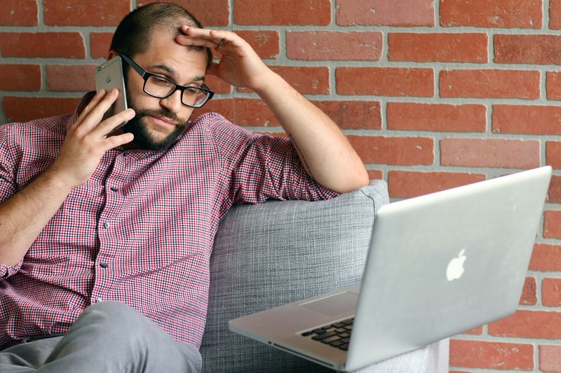 A man talking on the phone with a laptop in front of him. He has his hand to his head and looks irritated. 