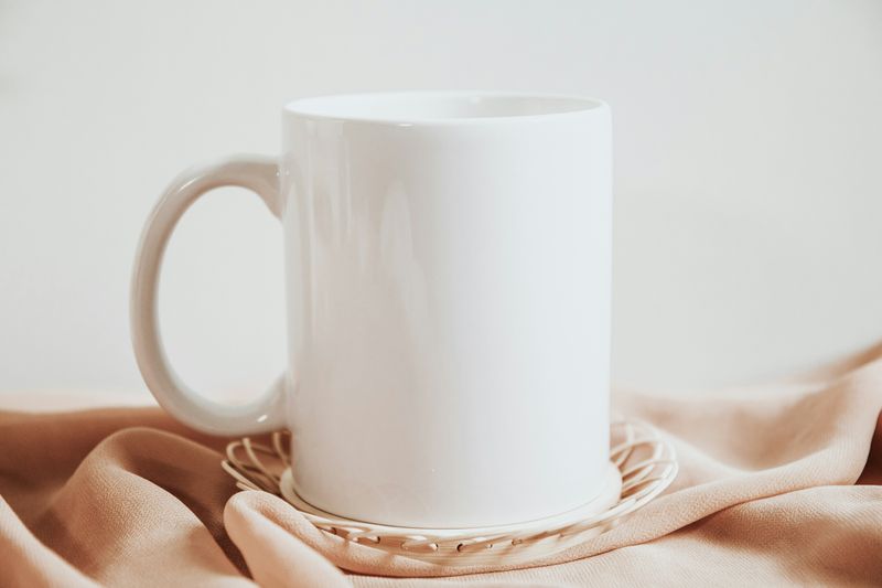 A white coffee mug sitting on a white doily on top of a beige blanket with a white background.