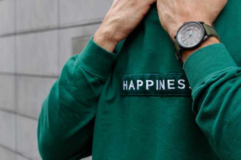 a man wearing a green fleece sweatshirt with the text 'HAPPINESS' on the sweatshirt, in front of a grey wall background