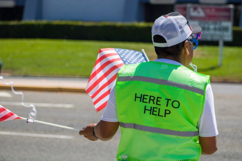 A man waving US flags wears a safety jacket that says, 'Here to help' on the back.