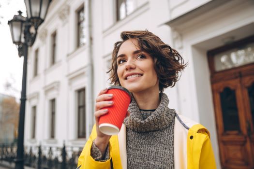 Young woman with brown hair wearing yellow rain coat, turtleneck sweater holding red Styrofoam-paper cup with plastic lid