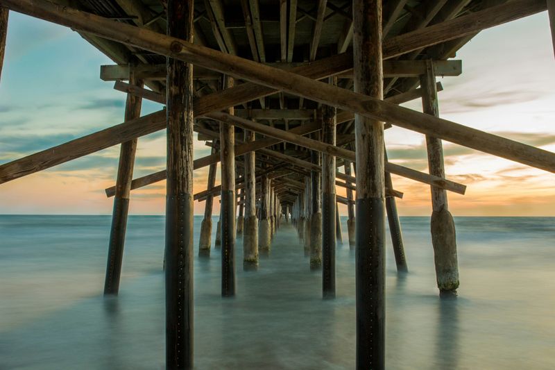 An point of view from beneath an ocean pier, with many support struts holding the pier up. 