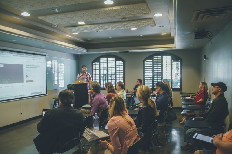 A student government committee meeting in a small university classroom. A speaker at the dais is presenting on a screen.