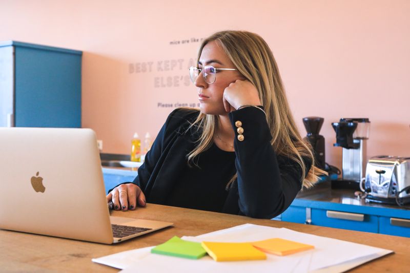 A woman with long, blonde hair sits in front of a laptop looking pensive.