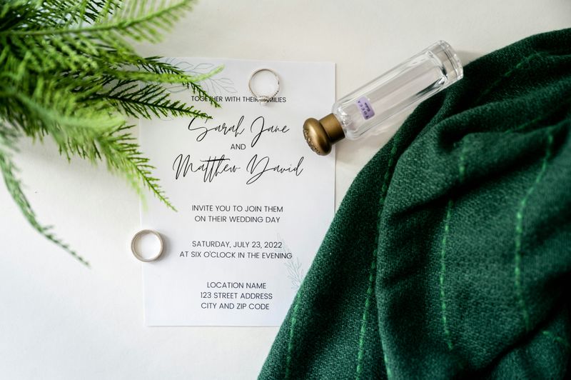 A photo of a wedding invitation surrounded by wedding rings, greenery, and perfume. 