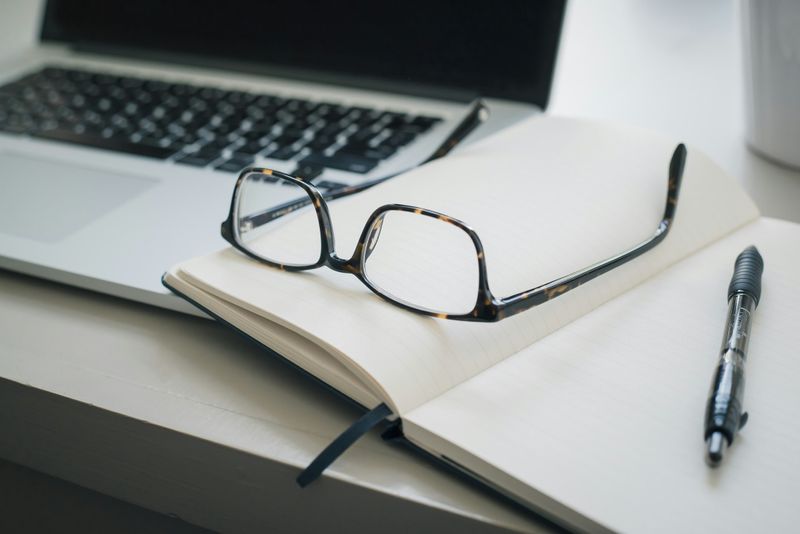 Upside-down reading glasses resting on an open notebook with a pen, with a laptop blurred in the background. 