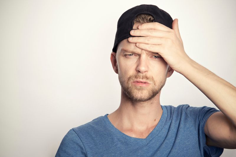 A man with backward black baseball cap touching forehead with an expression of pain.