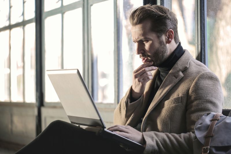 A man scratches his beard as he reads and processes information on his laptop. 