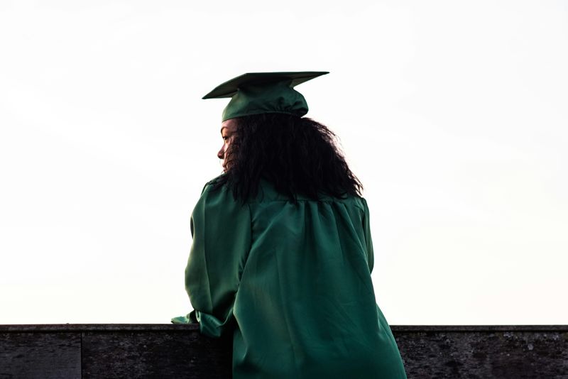 A graduate in a robe and gown looking into the distance while leaning against a wall.