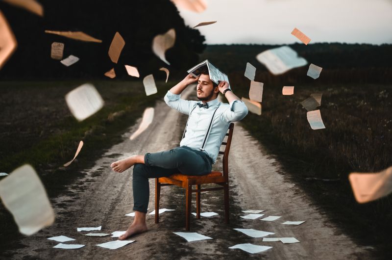 A man sits on a chair in the middle of the road. Papers fall on him from the sky.
