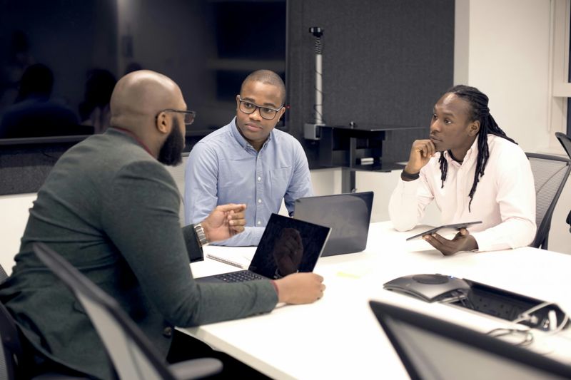 Three people on laptops and tablets at a table in an office, having a discussion.