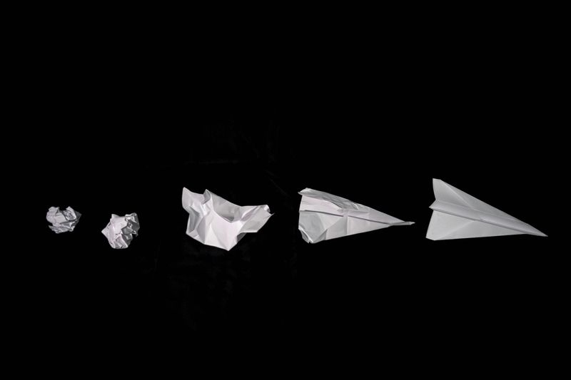 A crumpled paper gradually folding into a sleek paper airplane against a black background.