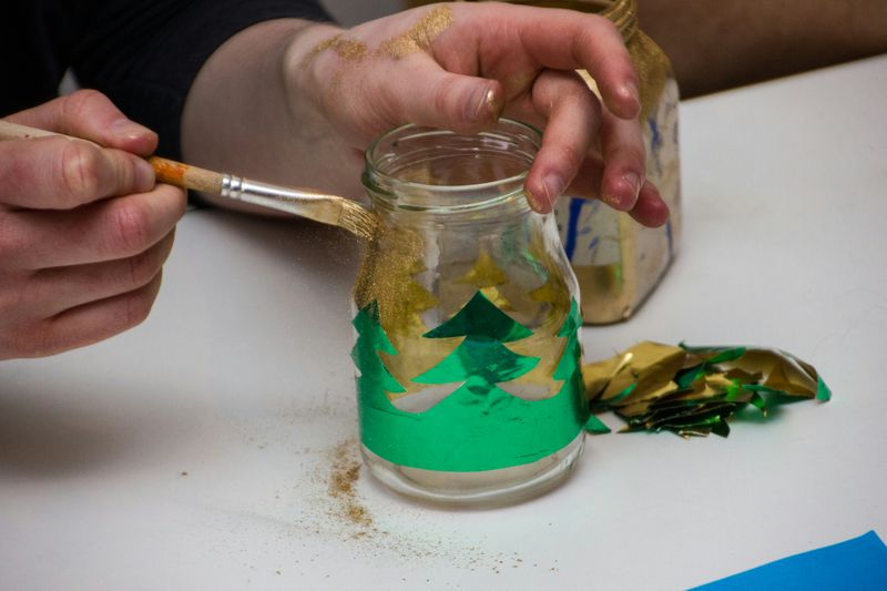 A hand painting gold paint on a glass jar with green tree decorations.