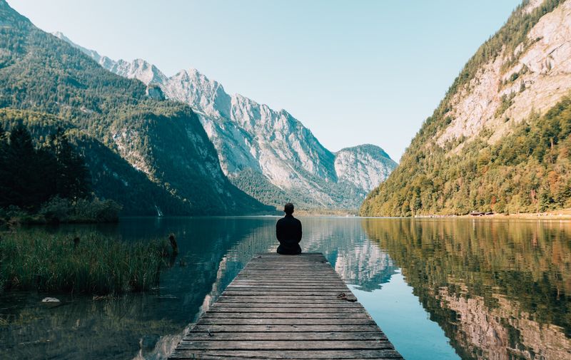 Person sitting on the edge of a dock overlooking the mountains and a lake.