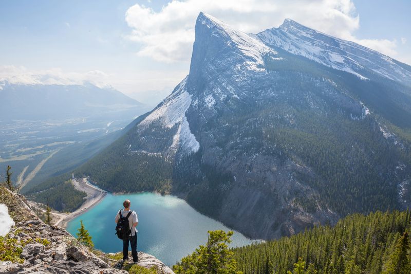 A person standing on a mountain peak overlooking a valley with a lake in the distance.