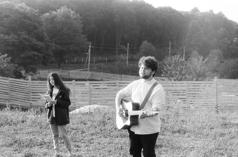 Man stands holding an acoustic guitar. Woman stands beside him holding sheet music (as if about to sing).