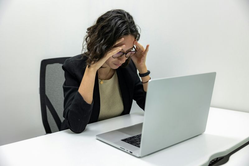A woman wearing glasses, leaning in and looking at the laptop on her desk, with both hands resting on her forehead.