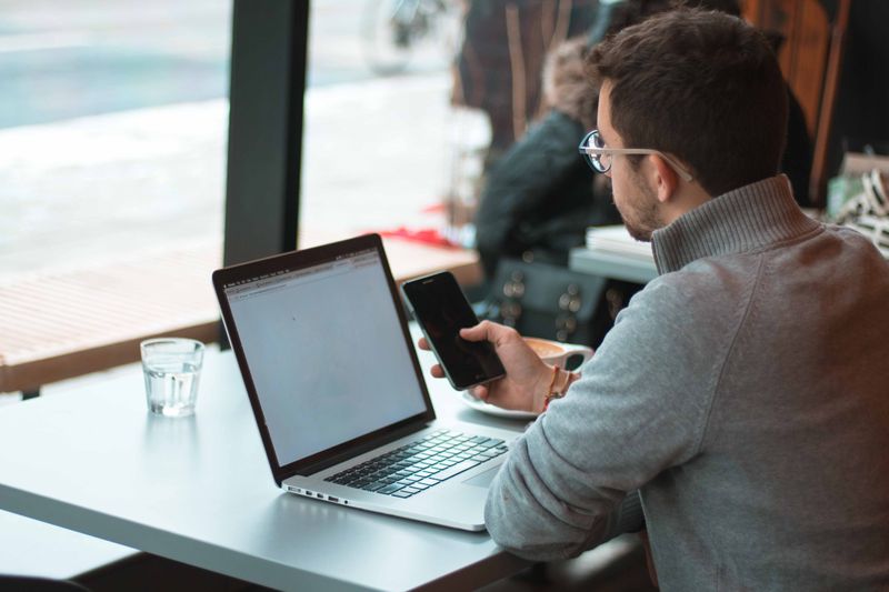 Man sits in front of laptop at table in restaurant, looking at smartphone